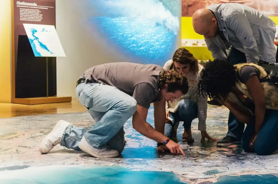 Visitors get up close and personal with a large panoramic floor map, searching for familiar places and landmarks. Geo-Stations on the exhibit floor feature quirky historic and contemporary place-based stories from around the Bay, such as the folk sculptures of the Emeryville mud flats, and the buried ships hidden under downtown San Francisco. Photo: Shaun Roberts. Courtesy of Oakland Museum of California.