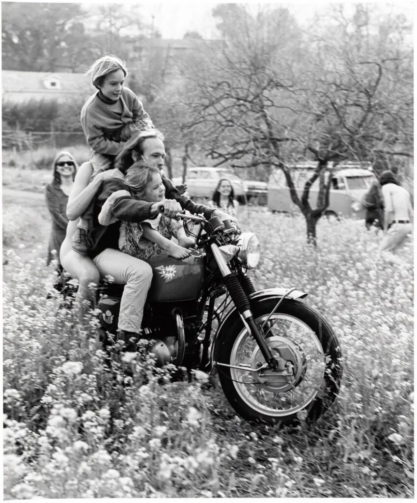 Black-and-white photograph of two adults and two children piled onto a motorcycle surrounded a meadow of flowers. The riders and motorcycle appear in the foreground, with a few smiling onlookers and a Volkswagen EuroVan in the background.