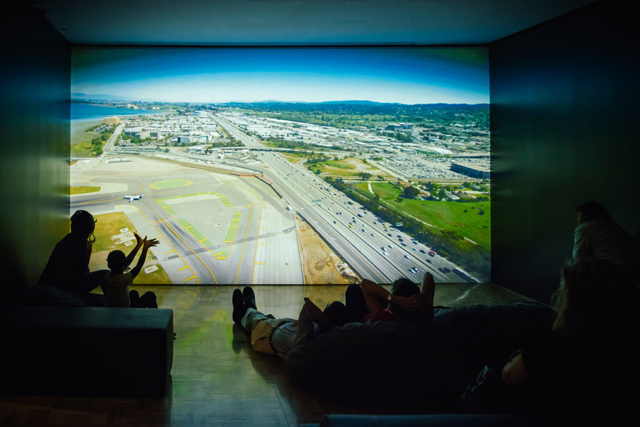 Visitors take an aerial tour of the shoreline of the San Francisco Bay Area, with a video projection commissioned by the Museum for its current exhibition, Above and Below: Stories From Our Changing Bay and created by the Center for Land Use Interpretation. The shore around the bay has been altered by humans and nature alike, and viewing it from above can consider the Bay's history and our relationship to our environment. Photo: Shaun Roberts. Courtesy of Oakland Museum of California.