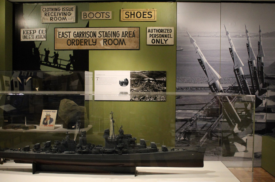 In the Military section of the gallery, the impact of major military development and expansion is demonstrated through historic objects and interactive media. A model of the USS Oakland, built at Hunters Point in San Francisco during World War II, rests in front of signs directing soldiers and recruits stationed on Angel Island and the Oakland Army Base. On the right is a photomural of Nike missiles, which were embedded in the mountaintop of Angel Island during the cold war, poised to defend the Bay against
