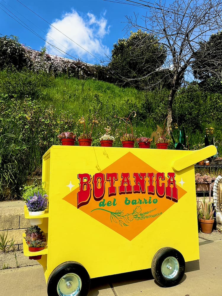 Photograph of a bright yellow cart, holding small potted plants, with the  text "Botanica del Barrio" in bold red and green letters